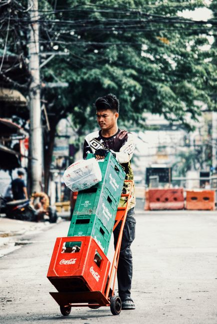 The image shows a young man with dark hair and casual clothing, standing outdoors on a street lined with trees and utility poles, during daylight. He is holding a trolley loaded with cases of Coca-Col