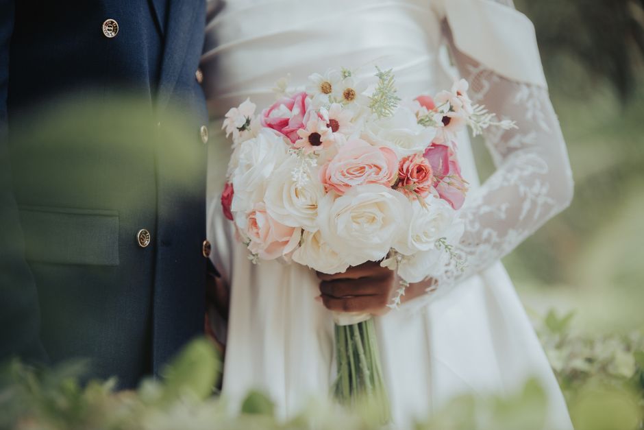 A bride and groom holding a floral bouquet with large white roses, soft pink roses, and smaller white and pink flowers, wrapped with a white ribbon. The bride is wearing a white lace wedding dress wit