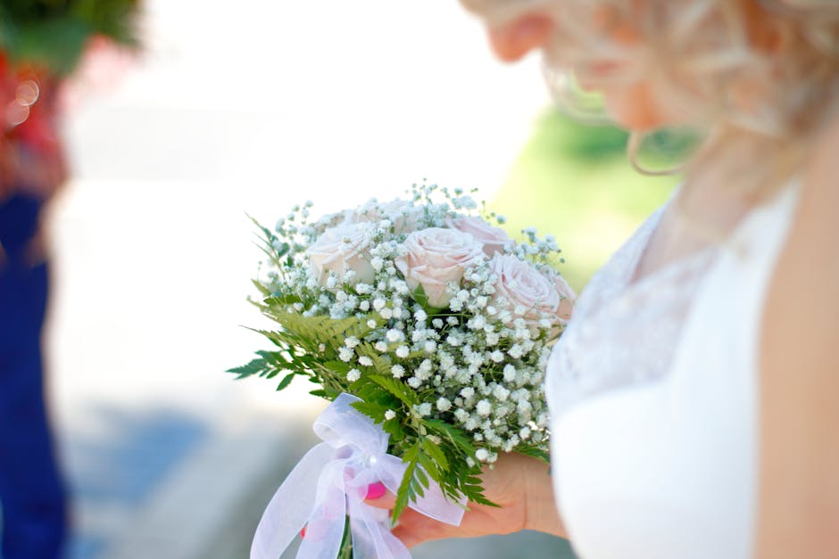 A person holding a floral bouquet featuring pale pink roses surrounded by small white baby’s breath and lush green foliage, wrapped with a light pink ribbon. The bouquet is arranged in a rounded, co