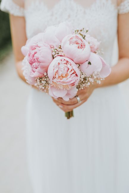 A close-up of a bride holding a delicate bouquet composed of large, soft pink peonies with layered petals, accented by small white gypsophila flowers and green stems. The bouquet is hand-tied with a s