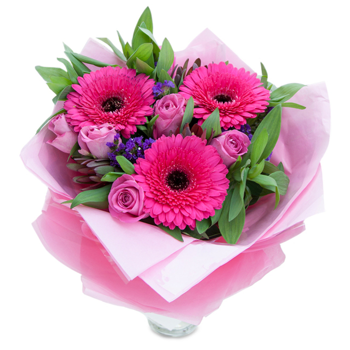 The composition centers on three bold gerbera daisies in a saturated fuchsia-pink, their concentric petals fanning outward and punctuated by a deep, almost-black eye that gives the bouquet a lively focal point. Soft, budding roses in a muted lavender-pink or dusty rose hue nestle close to the gerberas, their layered petals creating a gentle, velvety counterpoint and emitting a faint, pleasing rose scent. Interspersed throughout are sprigs of purple statice-small, clustered blooms that add texture and a cool violet contrast-while broad, glossy greens that resemble salal and eucalyptus form a verdant backdrop, introducing a crisp, slightly resinous fragrance and visual weight. The arrangement is finished in an inner wrap of soft pale pink tissue and an outer sheet of translucent fuchsia-pink cellophane with a subtle crinkle; the layered wrapping is folded around the stems with the attentive touch of a local florist, producing a neat, gift-ready presentation. At the lower edge the clear glass vase rim peeks through, signaling that the bouquet is destined for display and immediate enjoyment. Bathed in gentle daylight typical of a north-west London morning, the bouquet feels like a small celebration-bright, affectionate and composed with care-suitable for Mother's Day, birthdays, anniversaries or a cheerful delivery to a friend in Dollis Hill or a neighbour returning from a walk in Gladstone Park. It's the kind of arrangement that lifts a room with colour while promising to open further over the coming days.
