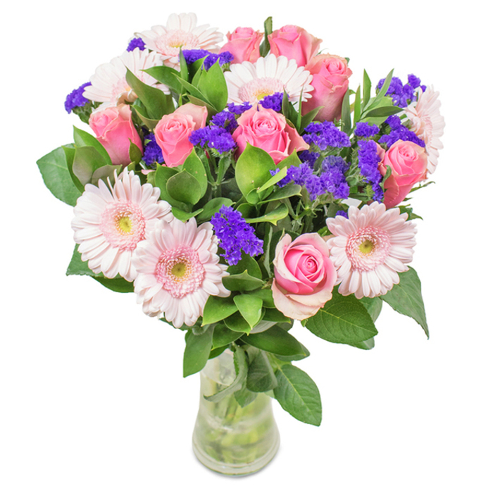 An elegantly balanced floral arrangement stands in a clear glass vase, set against a crisp white background-a reflection of Dollis Hill's timeless charm. The bouquet showcases about seven to nine medium pink roses, petals ranging from delicately furled to partially open, capturing the full spectrum of their natural beauty. Encircling the roses, five to seven pale pink Gerbera daisies stretch broad, soft petals around almost white centres, their tranquil hues reminiscent of peaceful park mornings in NW2. Clusters of vivid purple statice fill the spaces in between, adding both depth and playful vibrancy, while lush green foliage spills generously throughout, grounding the bouquet in the abundant greenery that Dollis Hill is known for. The arrangement sits in a slender glass vase where every stem and drop of water is visible, emphasizing the freshness of local delivery. Suitable for birthdays, anniversaries, or simply brightening a home or office near Dollis Hill Lane, this charming pink and purple bouquet is crafted to convey joy, celebration, and thoughtfulness in every detail-a loving reflection of the area's community spirit.